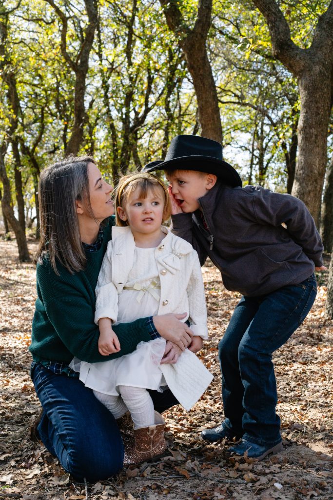 Mother kneeling with two young children outdoors in a wooded fall setting