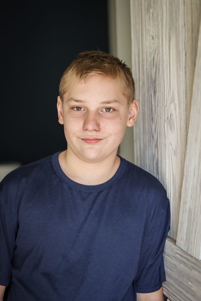 Young boy smiling at the camera while standing indoors near a wooden wall